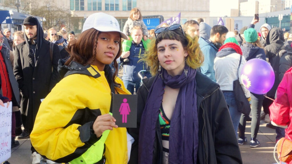 Two young women at a crowded outdoor rally; the left woman wears a yellow jacket and white cap, holding a sign with a pink female symbol